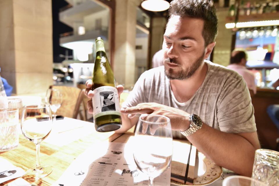 A man sitting at a table pointing at a bottle of wine.