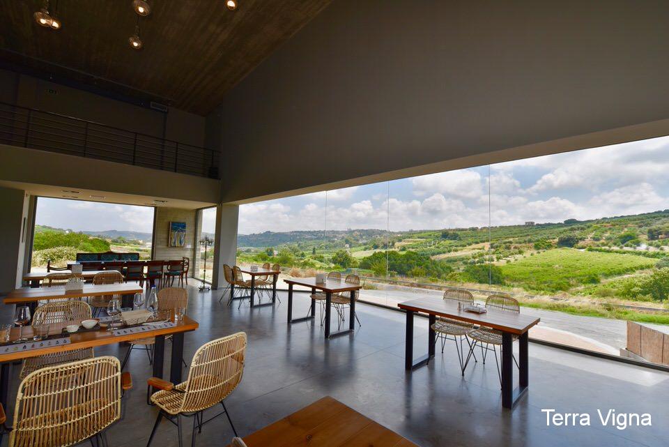 A room with tables and chairs with the view of wineries from the windows.