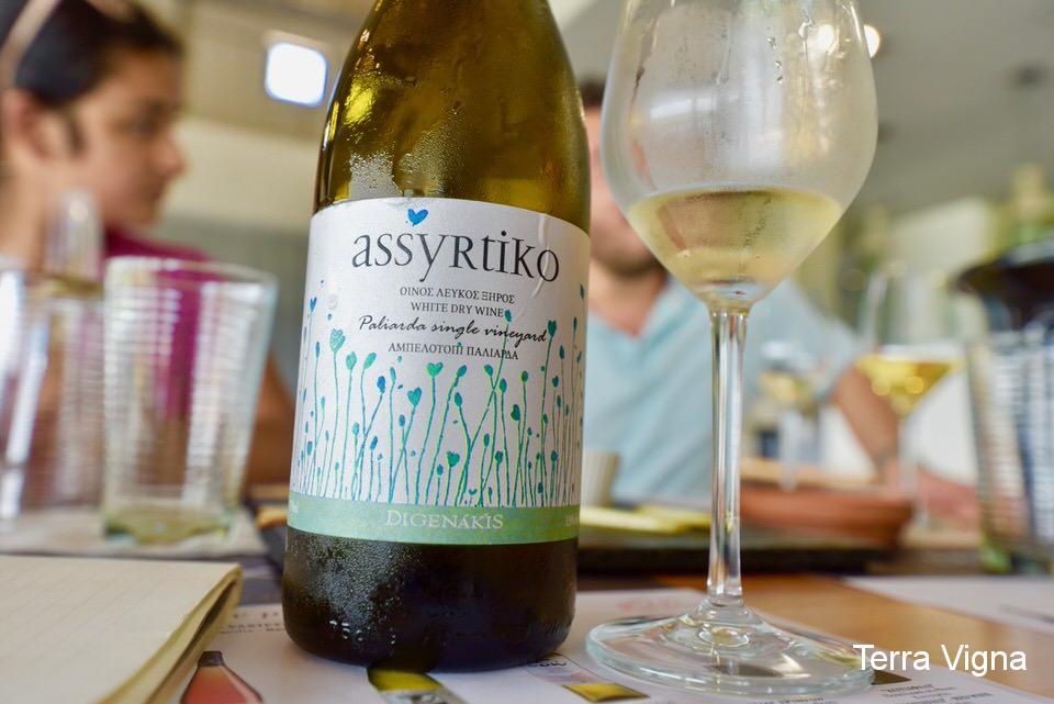 A bottle of white Greek wine on a table next to a glass of wine.